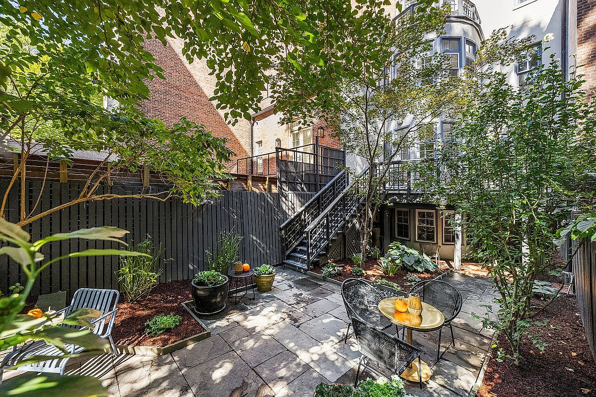 This image showcases a charming backyard garden area, featuring a stone patio with outdoor seating, including a table and chairs. The space is surrounded by lush greenery, including trees, bushes, and potted plants, creating a private and serene atmosphere. A dark-stained wooden fence and stairs leading to an upper level add architectural interest.