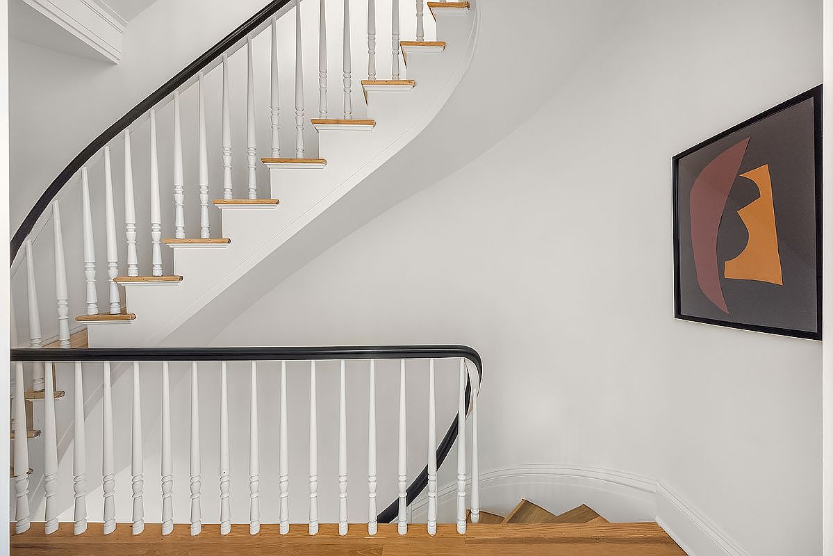 This image showcases a well-lit interior hallway featuring a staircase with white balusters and a dark handrail. The stairs have wooden treads, and a piece of modern art hangs on the wall, adding a touch of sophistication. The overall impression is clean, bright, and stylish.