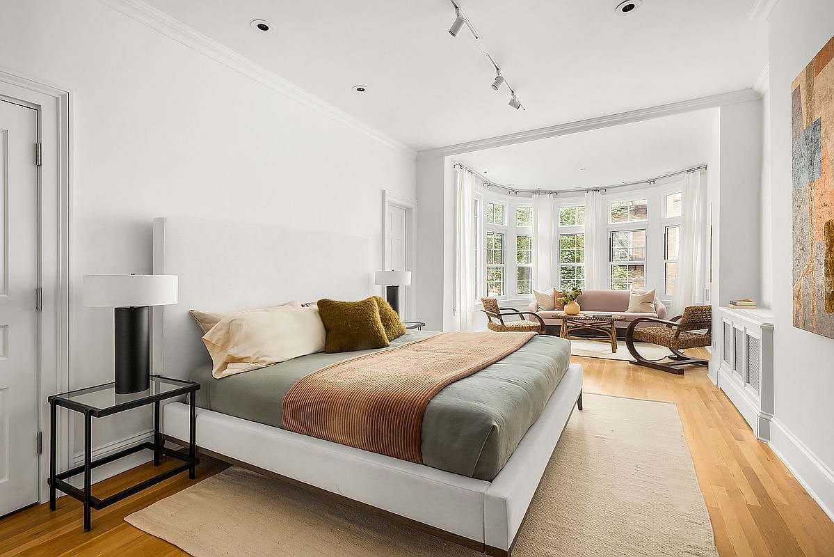 This is a bright and airy primary bedroom featuring a large bed with a white frame, neutral bedding, and a decorative throw. The room has hardwood floors, white walls, and a bay window area with seating, creating a relaxing and inviting atmosphere. A modern lamp sits on a glass-topped side table next to the bed.