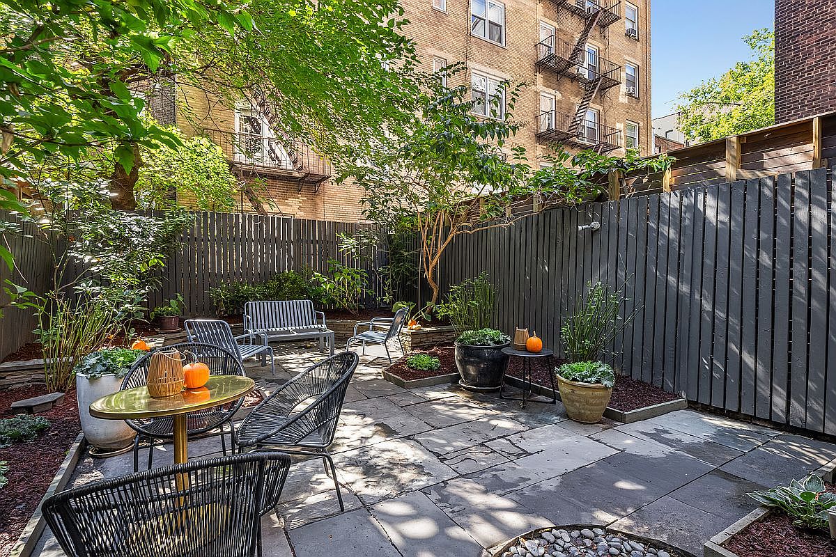 This is a charming backyard garden featuring a stone patio, a dark-stained wooden fence, and various seating arrangements including metal chairs and a bench. Potted plants and small trees add greenery and visual interest, while pumpkins provide a seasonal touch. The space is enclosed and private, offering a relaxing outdoor retreat.