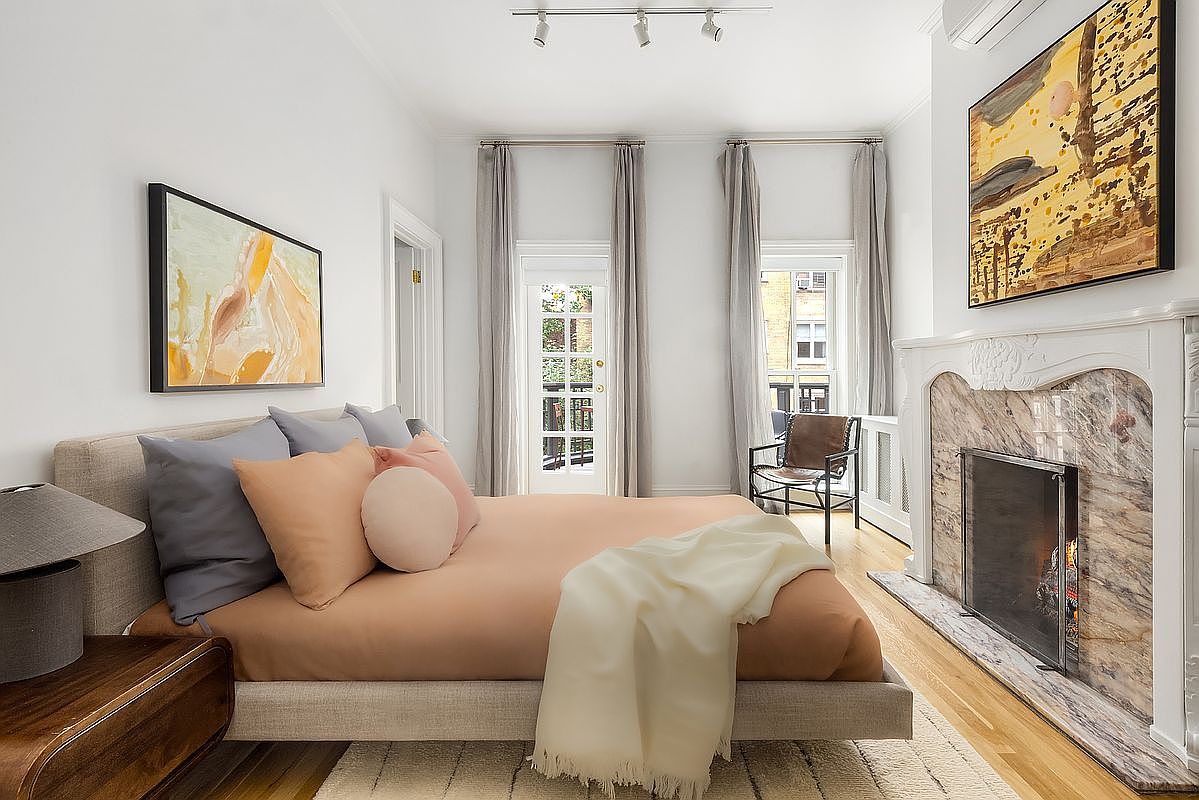 This is a well-lit primary bedroom featuring a neutral color palette with pops of color from the artwork and pillows. The room includes a bed with a light-colored frame, a decorative fireplace with marble accents, and access to a balcony through a glass door. The overall impression is one of comfort and understated elegance.