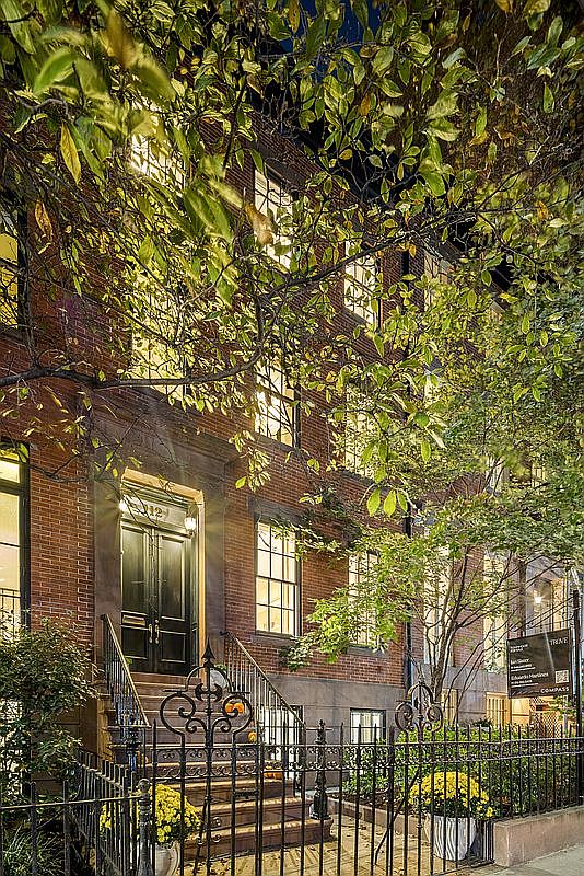 This is a front view of a charming brick townhouse. The entrance features a black double door with a decorative wrought iron railing leading up the steps. Lush greenery and trees frame the building, adding to its curb appeal and creating a welcoming atmosphere.
