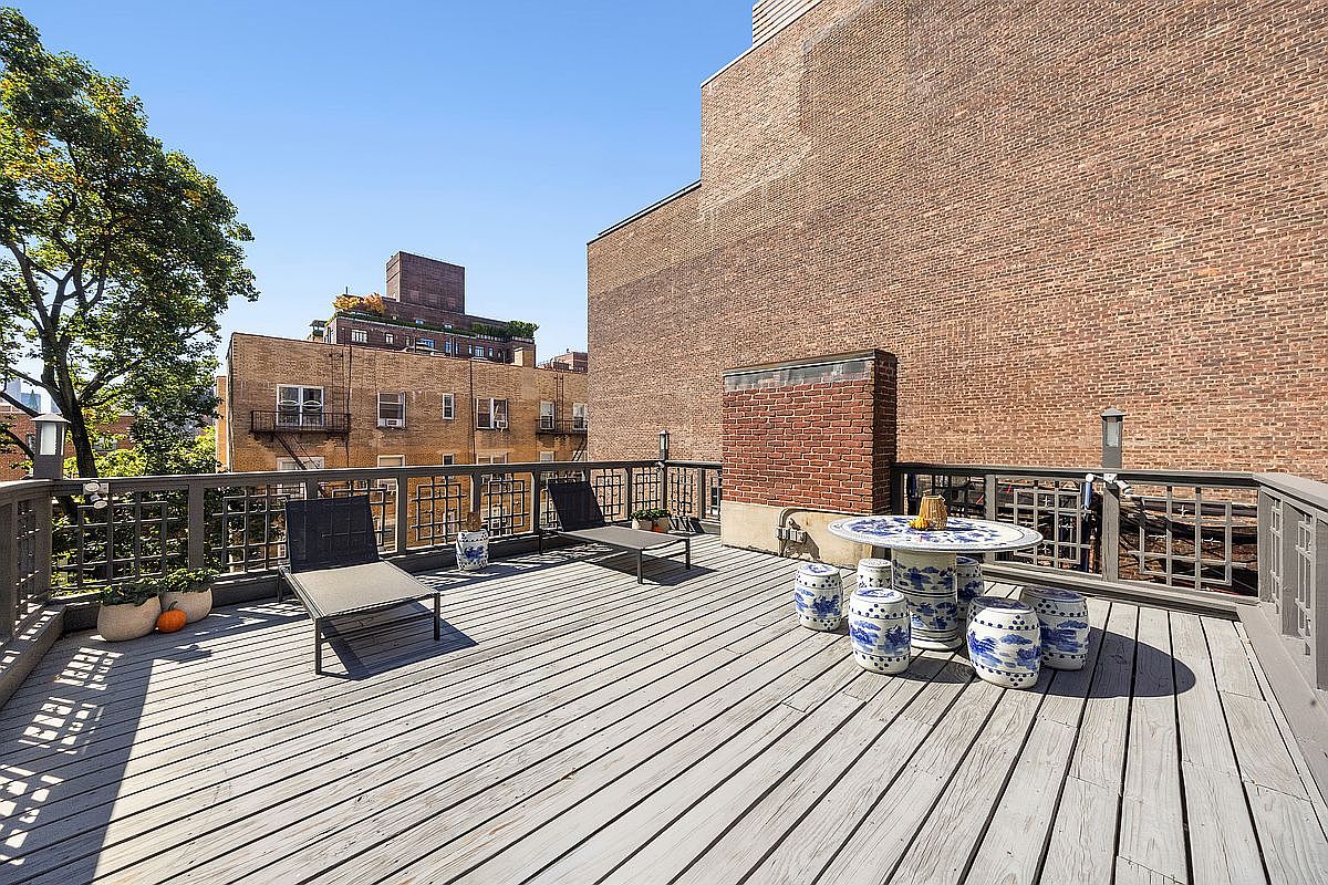 This image showcases a spacious rooftop deck with a wooden floor and a gray railing. The deck is furnished with lounge chairs and a unique outdoor dining set featuring a round table and blue and white ceramic stools. The backdrop includes brick buildings and a clear blue sky, creating an inviting outdoor living space.