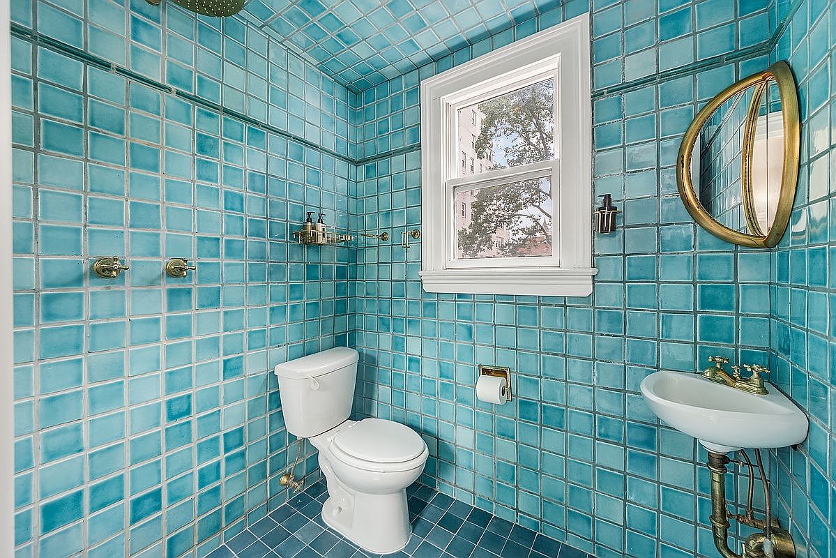 This bathroom features a unique design with blue square tiles covering the walls, floor, and ceiling, creating a cohesive and eye-catching aesthetic. The room includes a toilet, a wall-mounted sink with brass fixtures, and a round mirror, complemented by a window that provides natural light. The overall impression is a vintage-inspired, yet clean and well-maintained space.