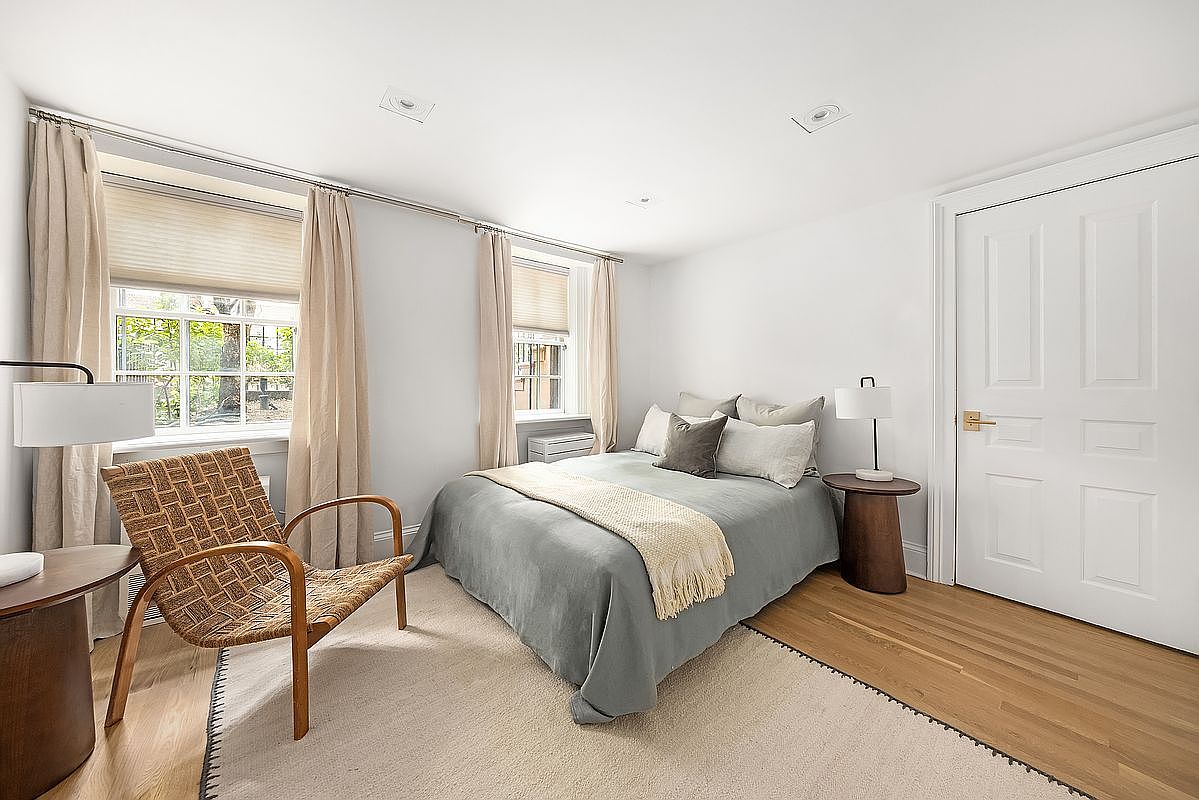 This is a cozy bedroom featuring a bed with a gray duvet and several pillows, flanked by two small side tables. A woven chair sits near the window, which is covered by blinds and curtains. The room has a neutral color palette, hardwood floors, and a simple, clean design, creating a serene and inviting atmosphere.