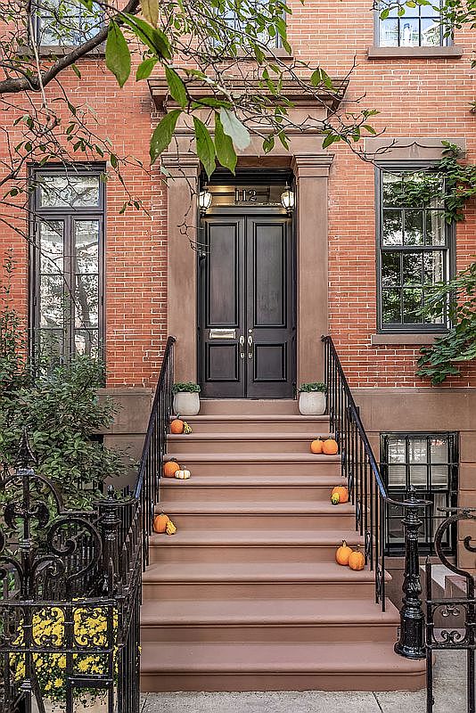 This is a charming entryway to a brick townhouse. The dark double doors are framed by a stone archway and flanked by sconce lighting. Steps lead up to the entrance, decorated with pumpkins, and wrought iron railings add a touch of elegance.