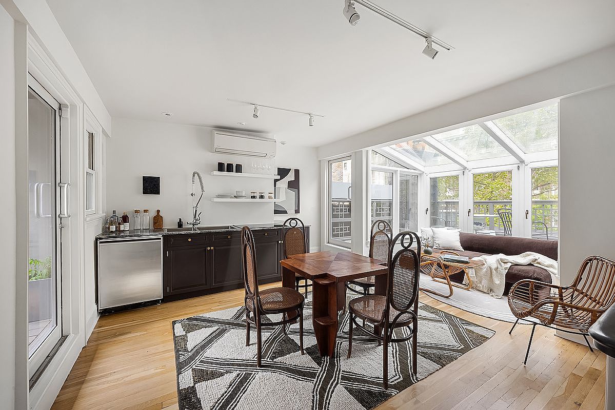 This interior shot showcases a dining area adjacent to a sunroom. The dining space features a square wooden table with four matching chairs, set upon a geometric patterned rug. A small kitchen area is visible to the left, and the sunroom offers ample natural light and a view of the outdoors, creating a bright and inviting atmosphere.