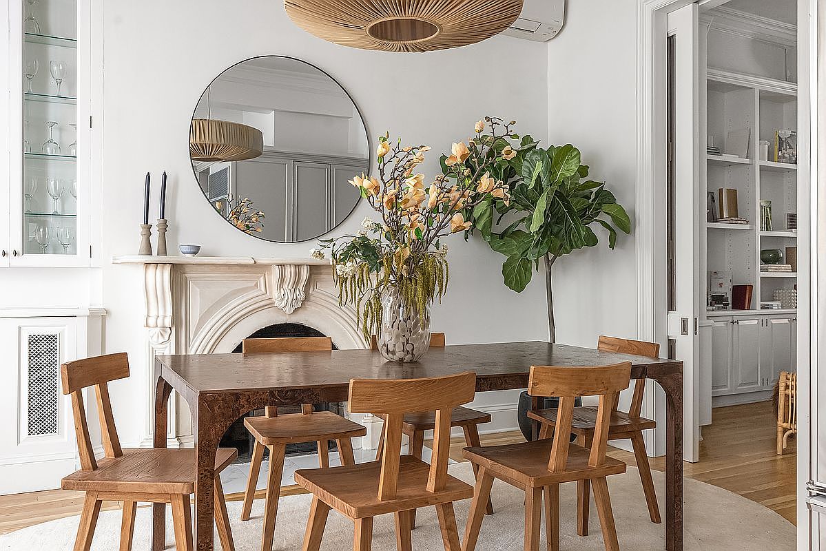 This is an interior shot of a dining room featuring a dark wood dining table surrounded by light wood chairs. A round mirror hangs above a decorative fireplace, and a large vase of flowers sits on the table. The room is well-lit and has a sophisticated, classic style.