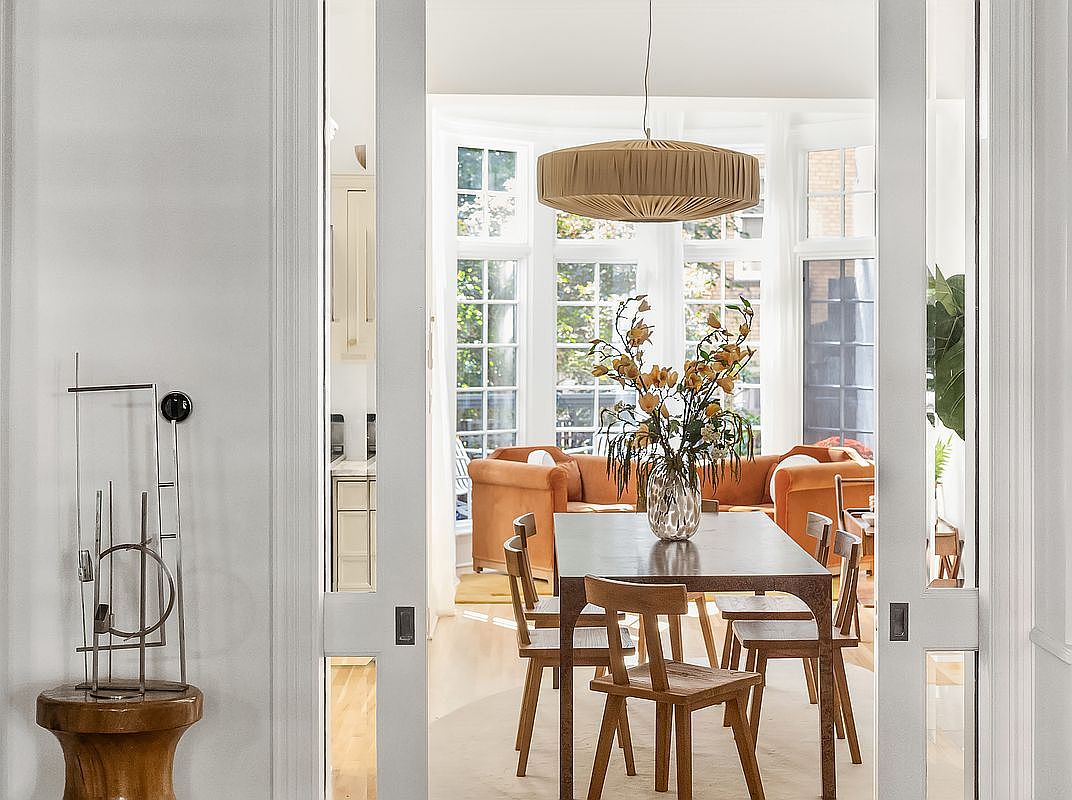 This image showcases a dining room with a view into a living area, framed by an open doorway. The dining room features a wooden table with matching chairs, a large floral arrangement, and a modern pendant light. The living area beyond includes comfortable seating and large windows, creating a bright and inviting atmosphere.