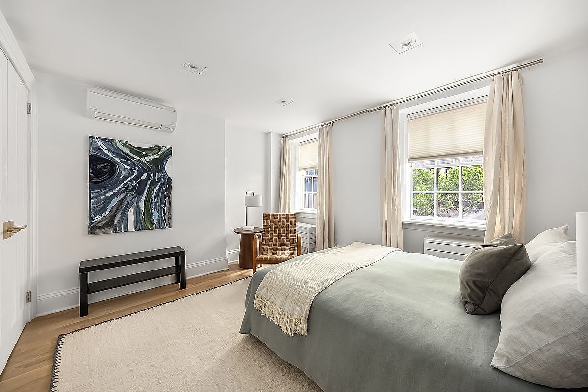 This is a bright and airy primary bedroom featuring a neutral color palette. The room includes a bed with light blue bedding and decorative pillows, a woven chair, and a dark wood bench. Natural light streams in through the windows, complemented by recessed lighting, creating a serene and inviting atmosphere.