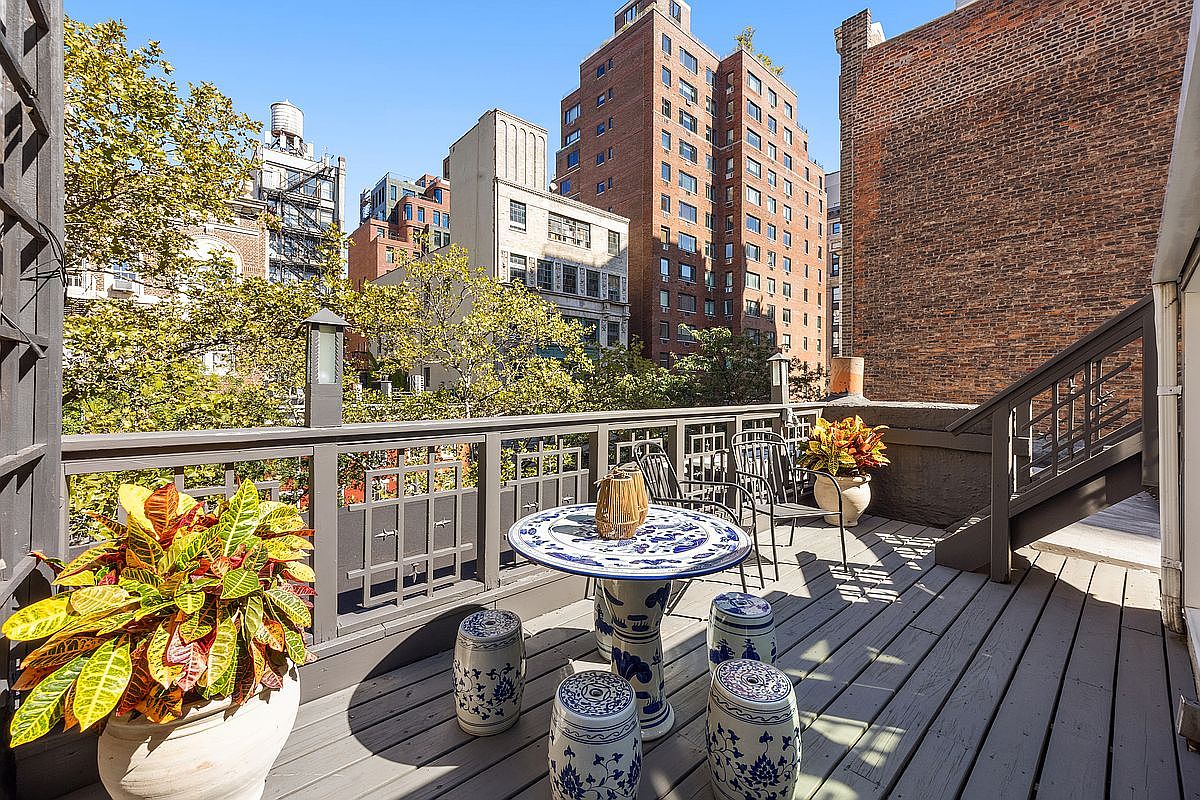 This image showcases a charming outdoor patio or deck area, furnished with a round table, chairs, and decorative stools, all set against a backdrop of urban buildings and greenery. The deck features a wooden floor and a railing, enhancing the outdoor living space. The presence of potted plants adds a touch of nature and color to the scene, creating an inviting and relaxing atmosphere.
