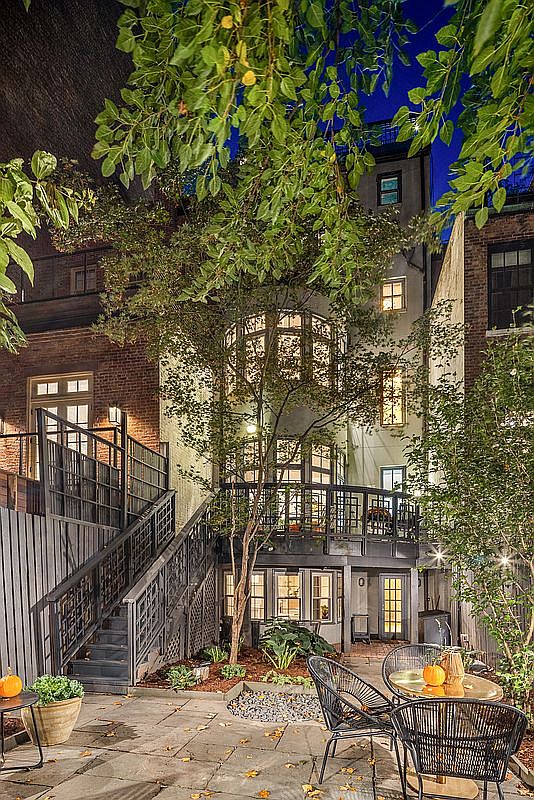 This image showcases a beautifully designed backyard garden at night, featuring a stone patio with outdoor seating, including wicker chairs and a round table with decorative pumpkins. A staircase leads to an upper level, and the building's architecture includes large windows and a balcony, creating an inviting and sophisticated outdoor living space.