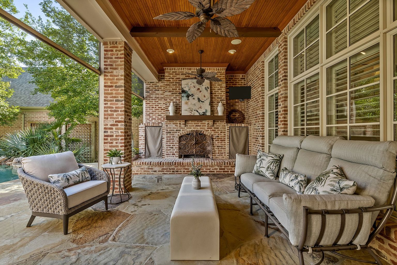 This image showcases a beautifully designed outdoor patio area. The space features comfortable seating arrangements, including a sofa and armchair, centered around a brick fireplace. The patio is covered with a wooden ceiling and stone flooring, creating a cozy and inviting atmosphere perfect for relaxation and entertaining.
