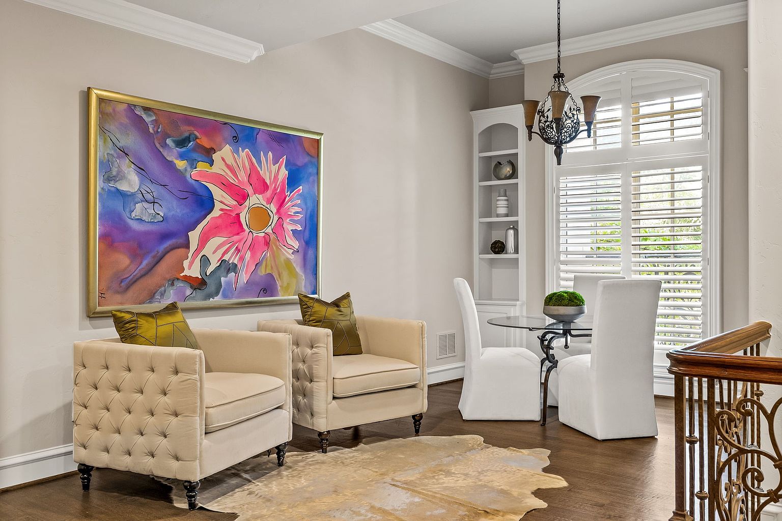 This dining room features a blend of modern and traditional elements. Two tufted armchairs sit beside a large, colorful painting, while a glass-topped table with white chairs is positioned near a window with shutters. A cowhide rug adds texture to the dark wood floor, creating a sophisticated and inviting space.