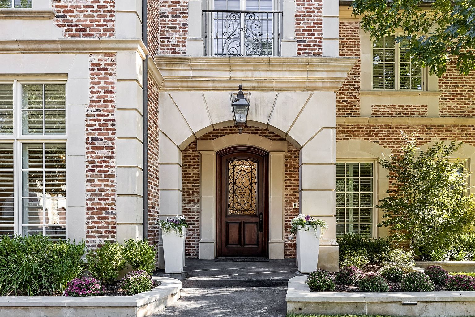 This is a front exterior shot of a brick home, focusing on the entryway. The arched doorway is framed by light stone columns and topped with a decorative lantern. Lush landscaping and potted plants flank the entrance, adding to the home's curb appeal and creating a welcoming atmosphere.