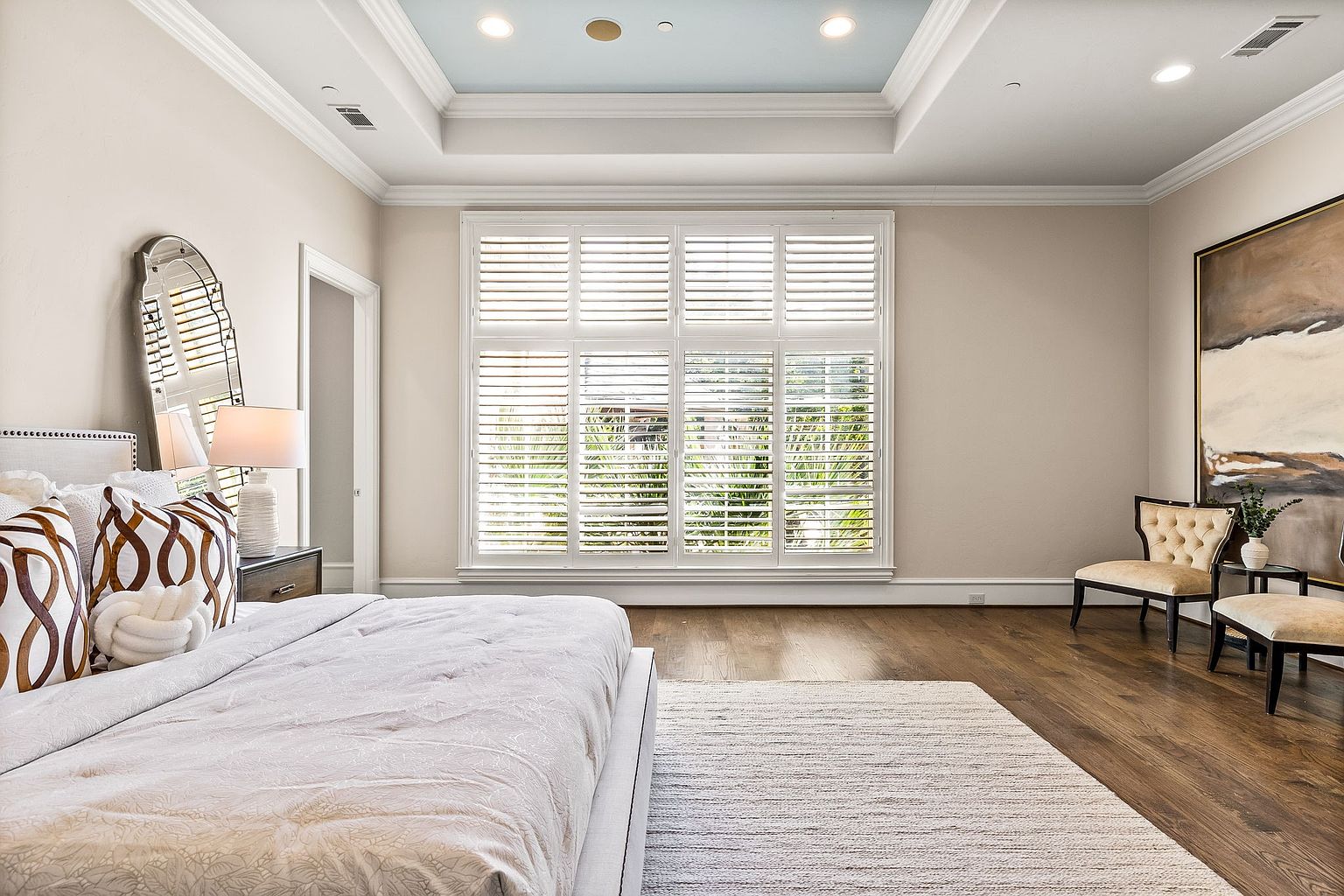 This is a bright and airy primary bedroom featuring a large window with plantation shutters, allowing natural light to fill the space. The room is decorated in neutral tones with a plush bed, decorative pillows, and a light-colored rug. Two chairs and a piece of artwork are placed in the corner, adding a touch of elegance to the room.