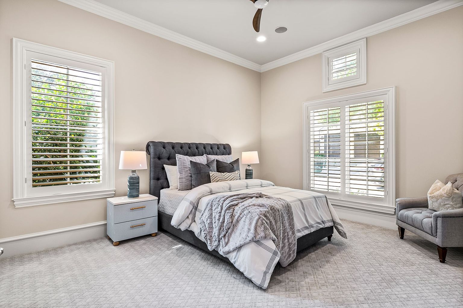 This is a well-lit primary bedroom featuring a tufted headboard bed with neutral-toned bedding and pillows. Two matching nightstands with lamps flank the bed. The room has large windows with white shutters, allowing natural light to fill the space, and a gray armchair sits in the corner, adding a touch of elegance.