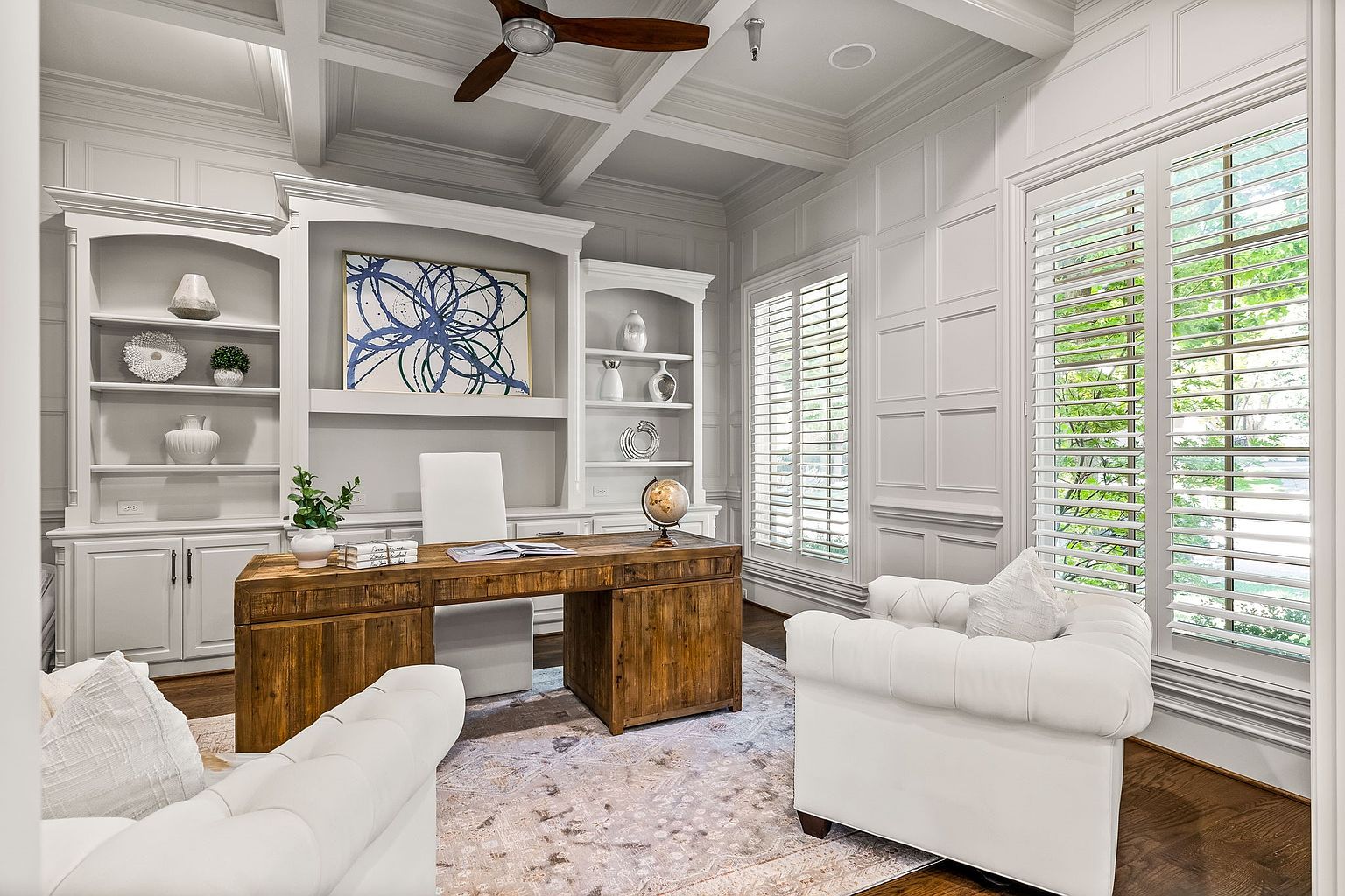 This is an interior shot of a well-appointed home office. The room features custom built-in shelving, a large wooden desk, and comfortable white armchairs. Natural light streams in through shuttered windows, creating a bright and inviting workspace.
