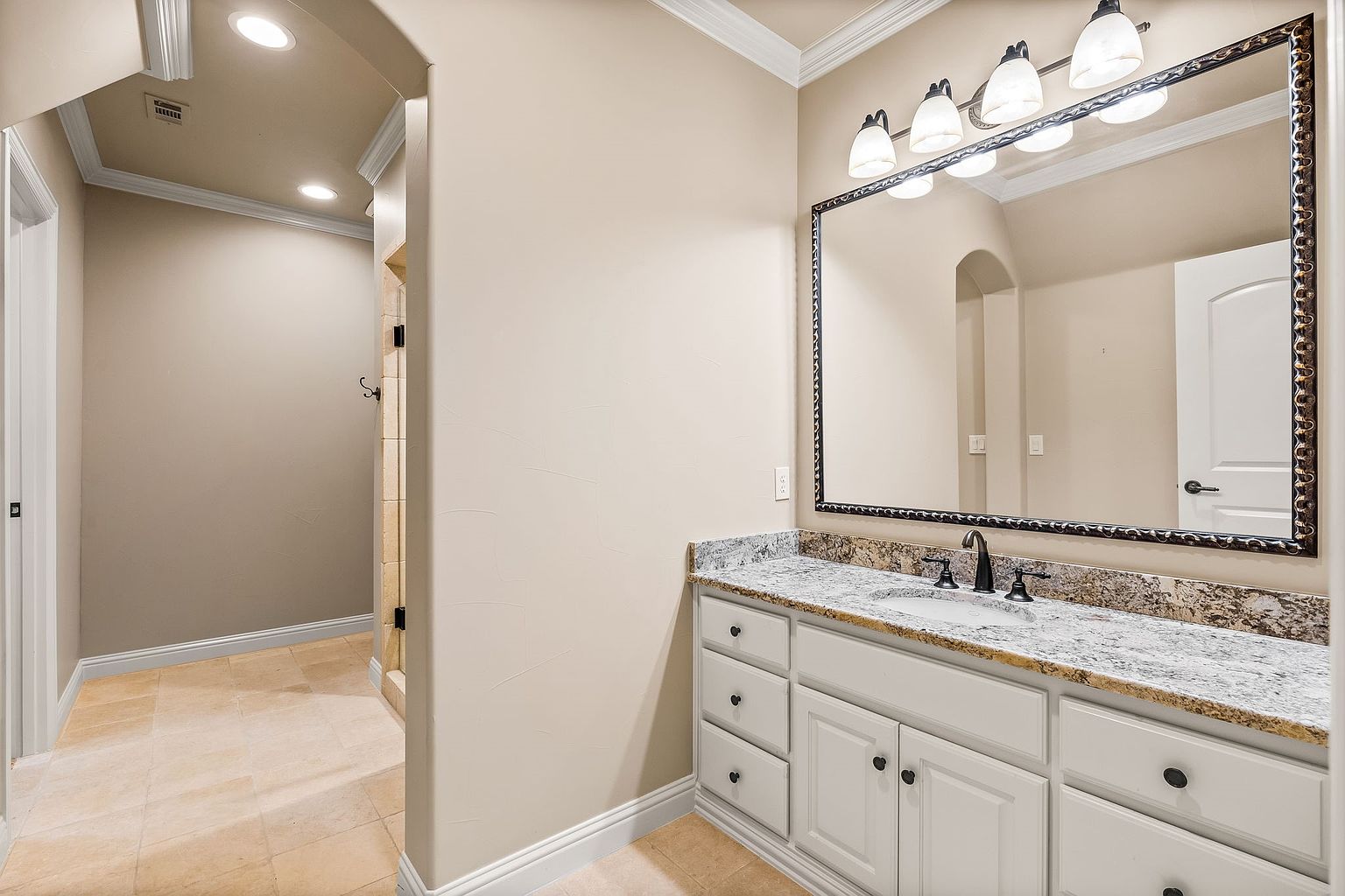 This is a well-lit primary bathroom featuring a vanity with white cabinetry and a granite countertop. A large, ornate mirror hangs above the sink, reflecting the room's neutral color palette and the adjacent hallway. The flooring appears to be tile, and the overall impression is clean and elegant.