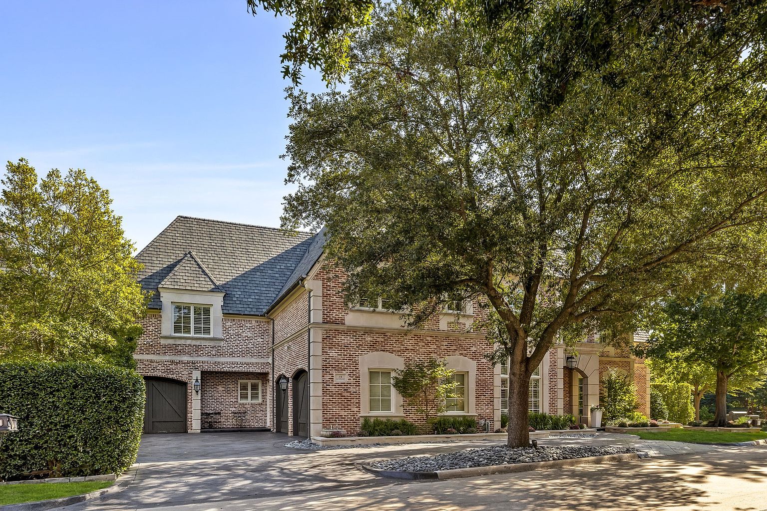 This is a front exterior view of a stately brick home with a dark gray roof and elegant architectural details. A large, mature tree dominates the foreground, casting dappled shadows on the paved driveway and manicured landscaping. The home features multiple windows with light-colored trim and a covered entryway, exuding a sense of timeless elegance and curb appeal.