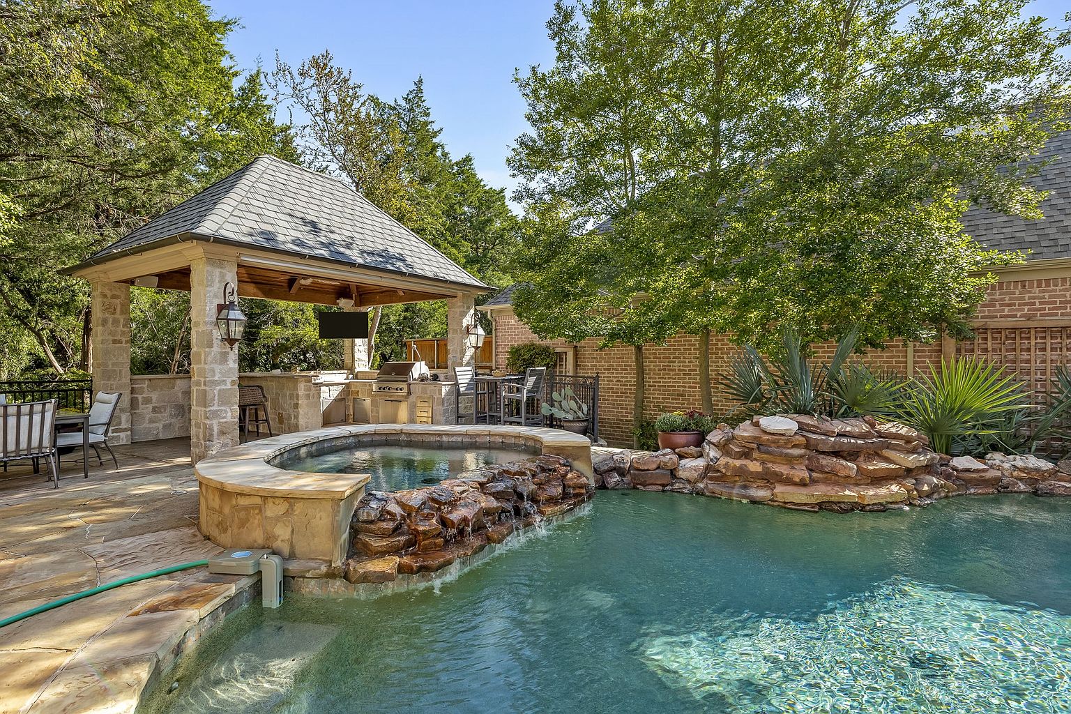 This image showcases a luxurious backyard pool and spa area. The spa features a stone waterfall cascading into the pool, surrounded by lush landscaping and mature trees. A covered outdoor kitchen and bar area with a built-in grill and seating enhance the entertainment possibilities, creating an inviting and upscale outdoor living space.