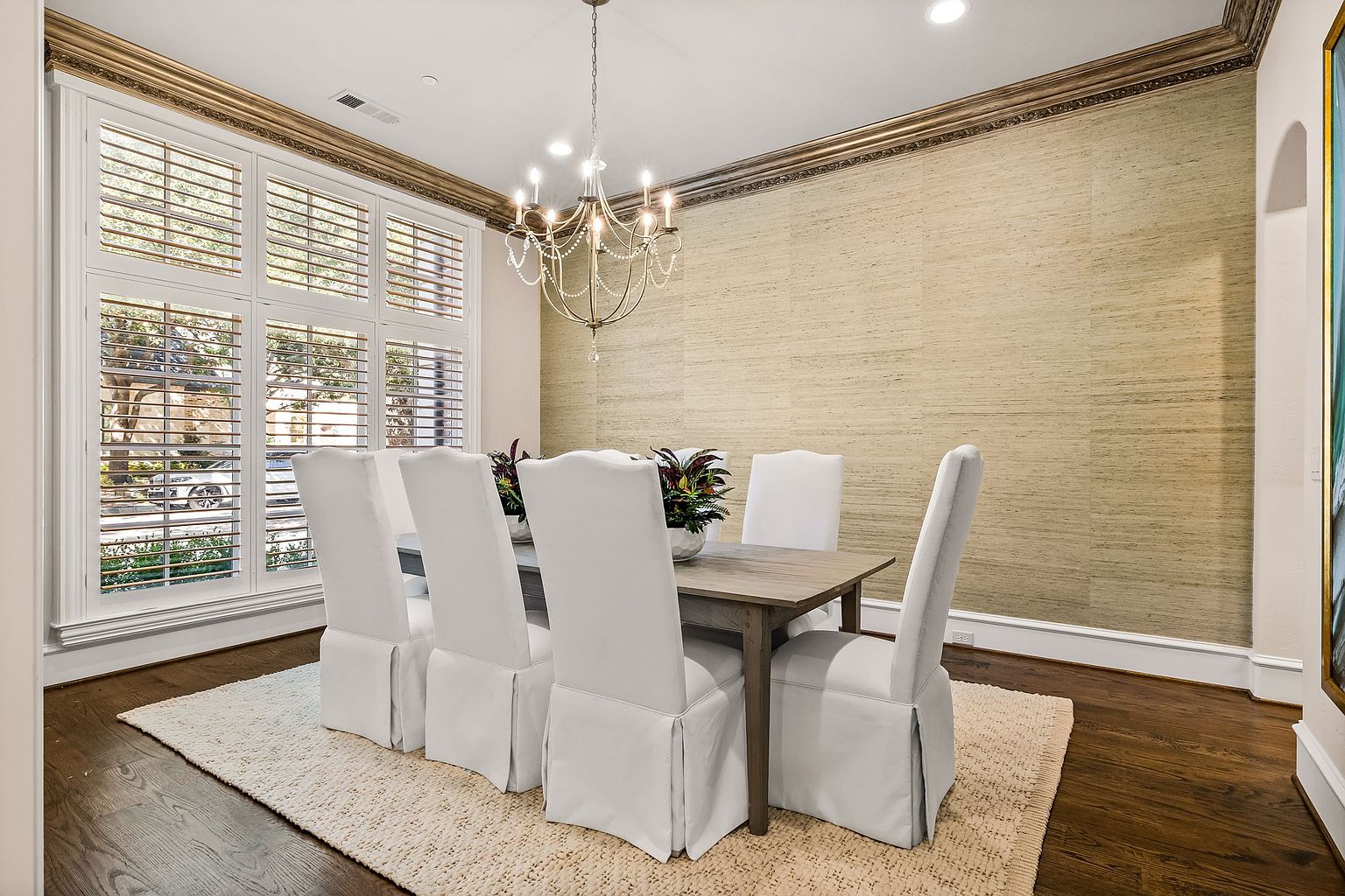This is an interior shot of a dining room featuring a wooden table with six white upholstered chairs. A chandelier hangs above the table, and a large window with shutters provides natural light. The room is decorated with neutral-toned wallpaper and dark hardwood floors, creating an elegant and inviting atmosphere.