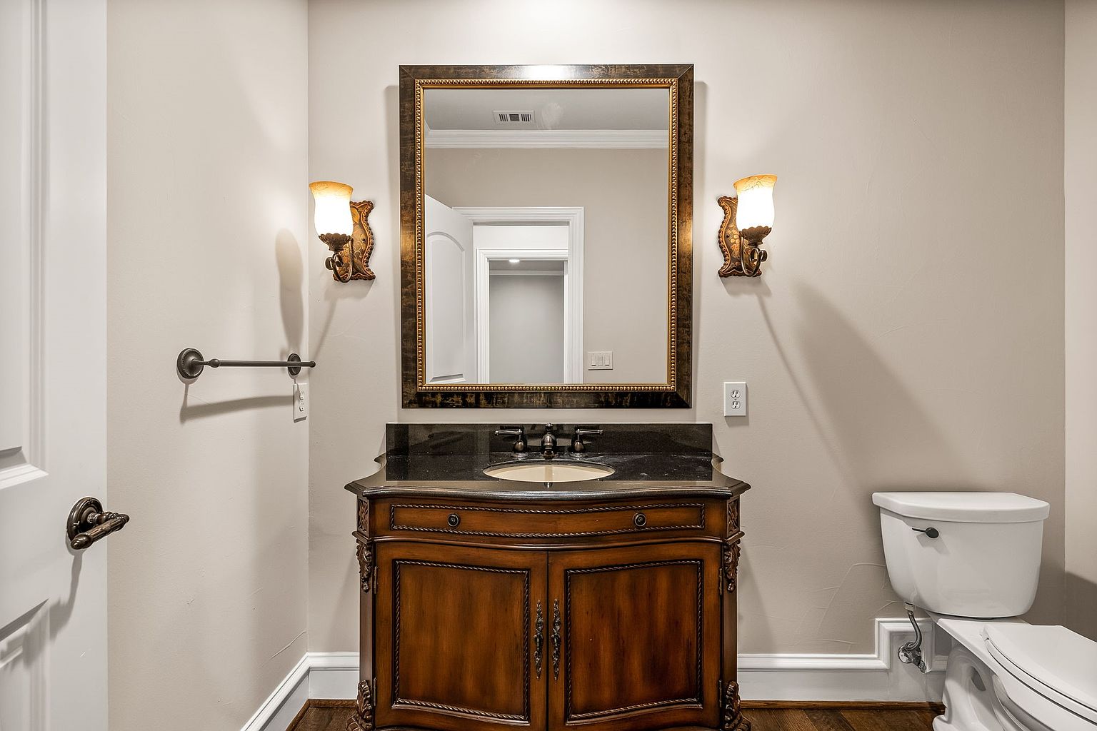 This is a well-appointed guest bathroom featuring a dark wood vanity with a black countertop and a decorative framed mirror. The walls are painted in a neutral tone, and sconces flank the mirror, providing warm lighting. A toilet is visible to the right, and a towel bar is on the left wall, creating a functional and stylish space.