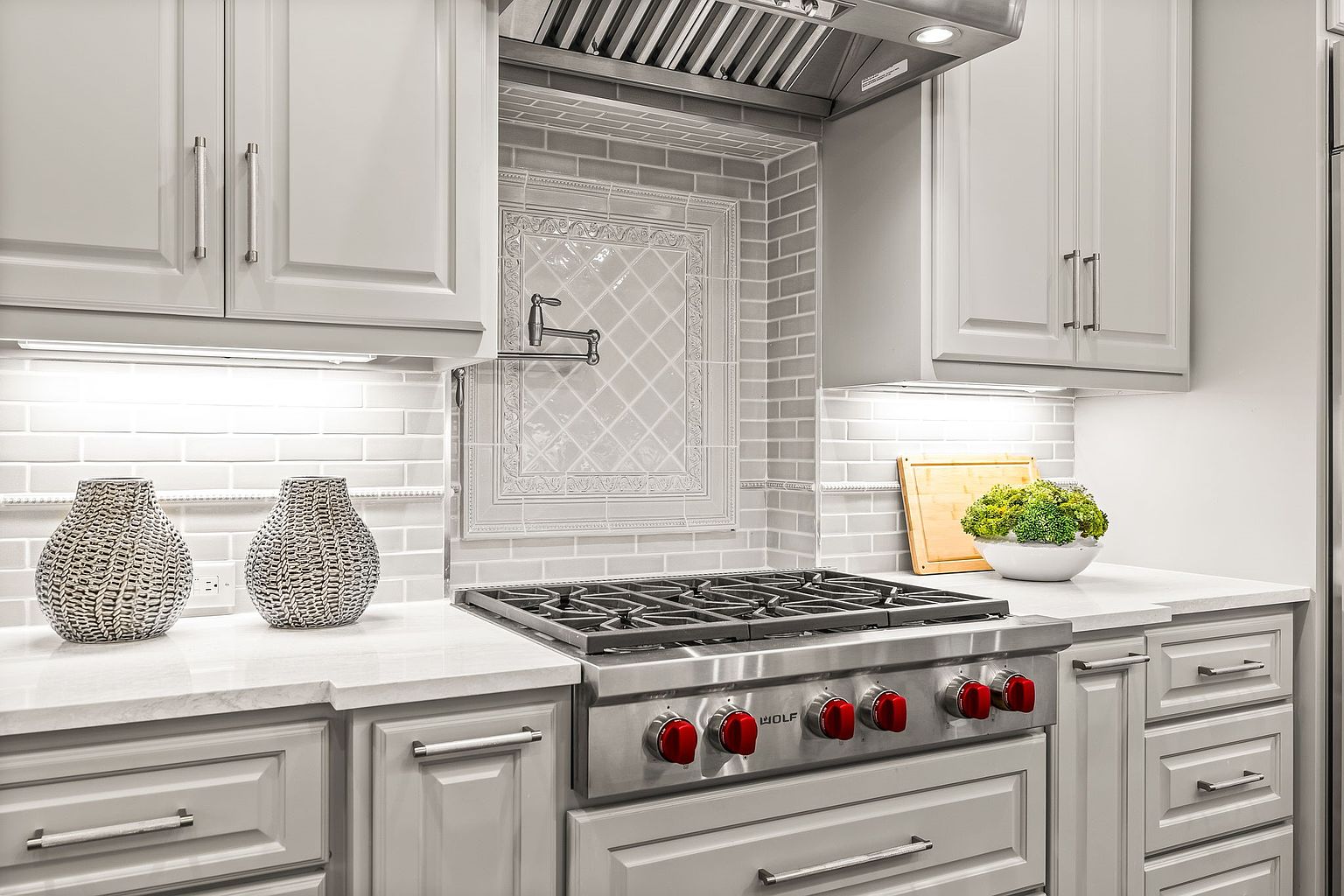 This is a well-lit kitchen featuring gray cabinetry with silver hardware and a stainless steel Wolf range with red knobs. The backsplash is a light gray subway tile with a decorative inset above the range. The countertops are a light-colored stone, and decorative vases and a plant add a touch of elegance.