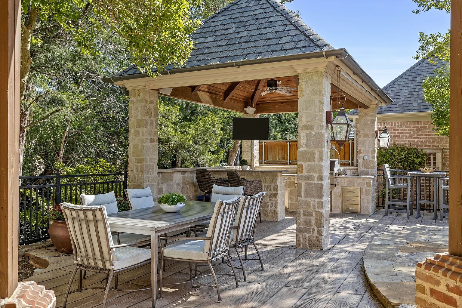 This image showcases a luxurious outdoor patio area, featuring a stone-covered pavilion with a built-in outdoor kitchen and bar. The patio is furnished with a dining table and chairs, creating an inviting space for entertaining. The stone construction and elegant design elements contribute to a high-end and sophisticated outdoor living experience.