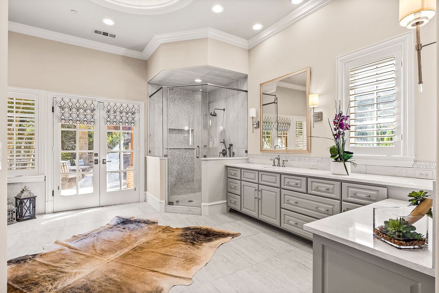 This is a luxurious primary bathroom featuring a glass-enclosed shower with gray tile, a double vanity with gray cabinets and white countertops, and a large mirror. The room is well-lit with natural light from a window with shutters and artificial lighting from sconces and recessed lights. A decorative cowhide rug adds a touch of warmth to the tile floor.