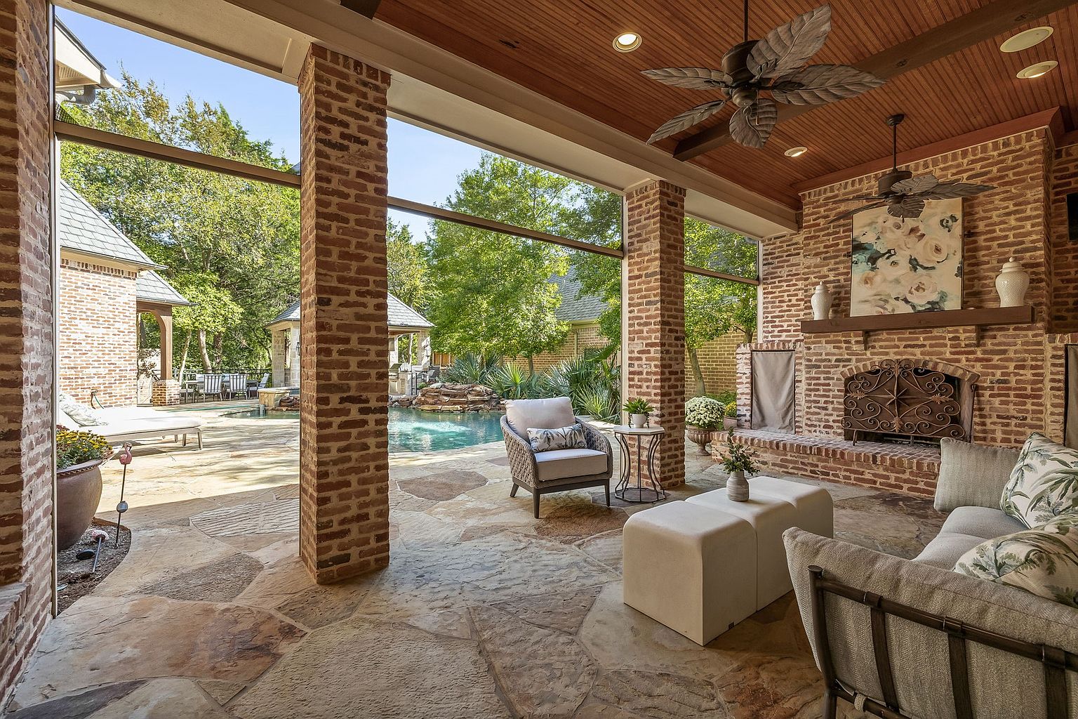 This image showcases a luxurious outdoor patio area, featuring brick columns and a ceiling with wooden planks and a ceiling fan. The patio includes comfortable seating arrangements, such as a sofa, armchairs, and ottomans, arranged around a brick fireplace. The flooring is made of natural stone, and the patio overlooks a swimming pool and landscaped garden, creating a serene and inviting atmosphere.