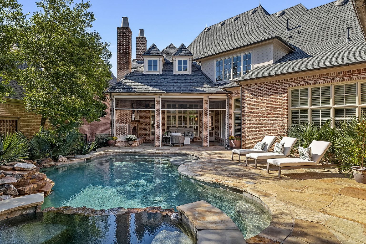 This image showcases a luxurious backyard pool and spa area. The pool features a natural stone waterfall and is surrounded by a stone patio with lounge chairs. The architecture of the house is visible in the background, featuring brick and stone elements, dormer windows, and a covered patio area, creating an inviting outdoor living space.