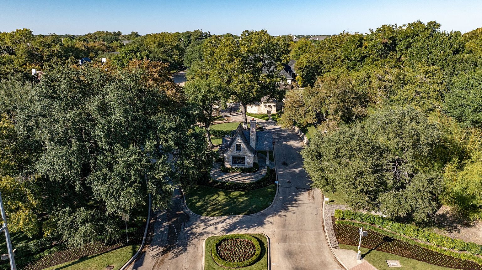 This aerial shot showcases a charming stone house nestled among lush trees, creating a sense of privacy and tranquility. The property features a well-manicured lawn and a circular driveway, adding to its curb appeal. The surrounding greenery and mature trees enhance the overall aesthetic, suggesting a peaceful and secluded setting.
