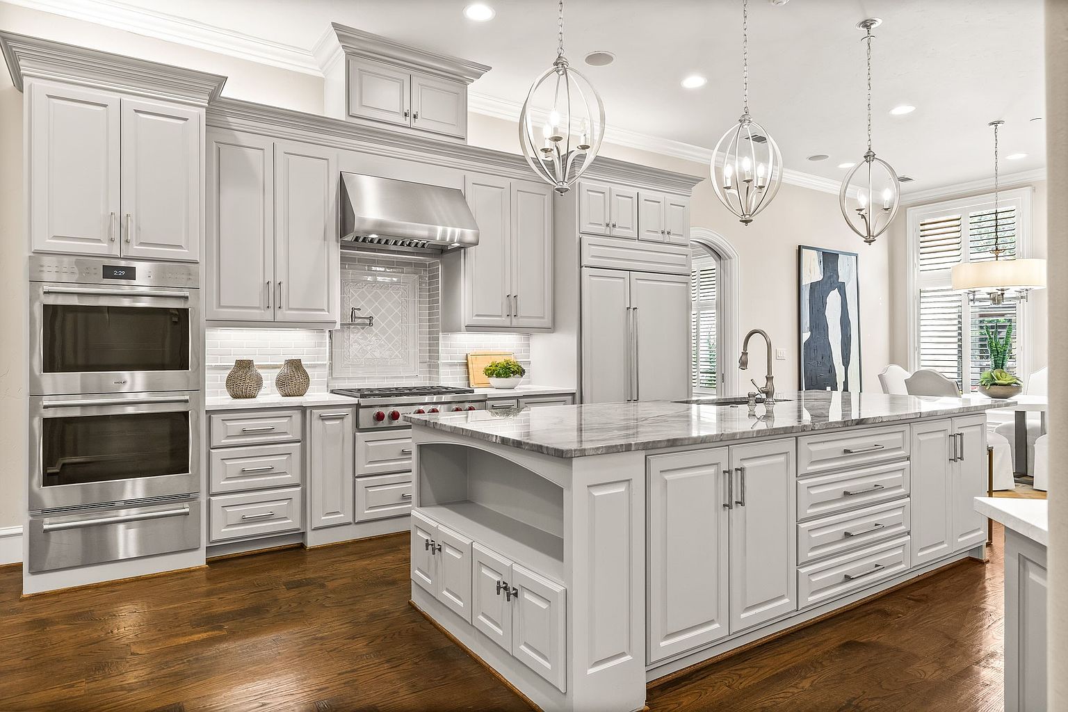 This is a well-lit, modern kitchen featuring gray cabinetry, stainless steel appliances, and a large island with a marble countertop. The kitchen includes a double oven, a gas range with a stainless steel hood, and a built-in refrigerator. Three globe pendant lights hang above the island, and the dark hardwood floors add warmth to the space. The perspective is from a medium distance, capturing the entire kitchen area.