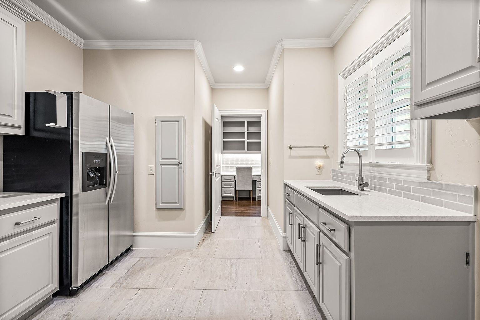 This is an interior shot of a pantry area, featuring a stainless steel refrigerator and white cabinetry. The pantry includes a sink and countertop area, with a view into a small office space through an open doorway. The flooring is light-colored tile, and the overall impression is clean and organized.