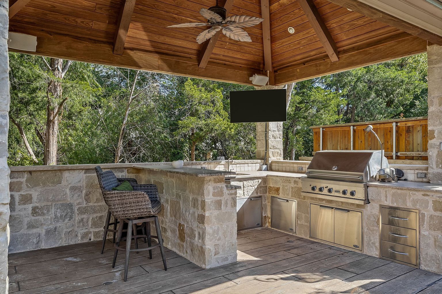 This image showcases an outdoor kitchen and bar area under a wooden gazebo. The space features stone countertops, stainless steel appliances including a grill and storage, and bar seating. The area is surrounded by lush greenery, creating a private and inviting outdoor living space.