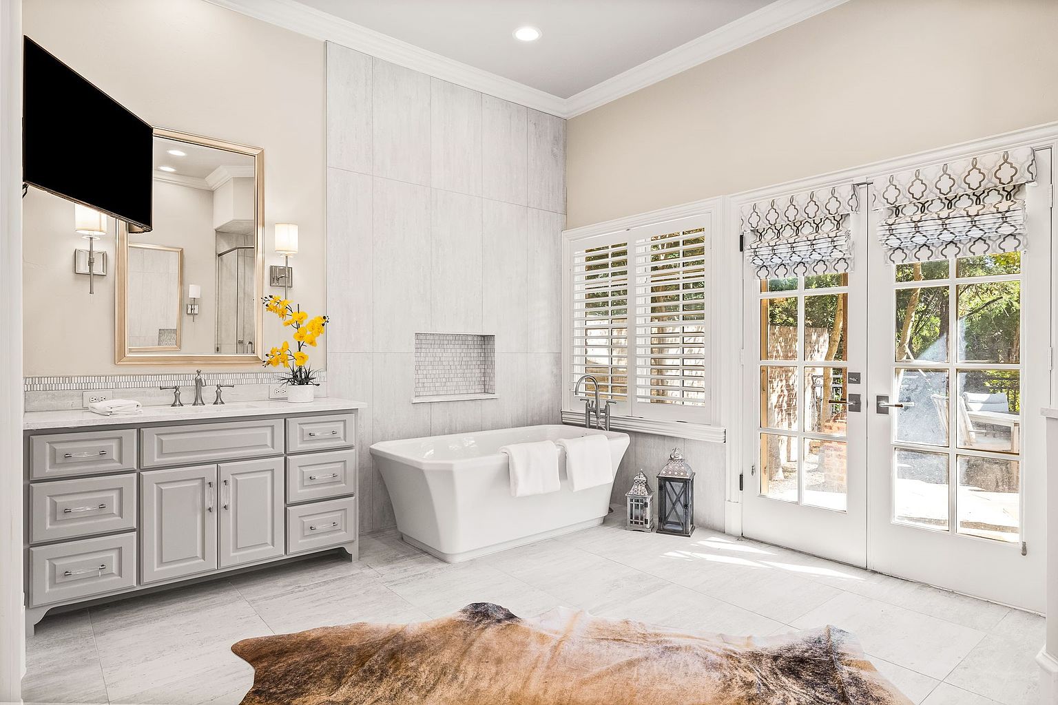 This is a bright and spacious primary bathroom featuring a large vanity with ample storage, a freestanding bathtub, and a separate tiled shower area. The room is well-lit with natural light streaming through the windows and glass doors, which are adorned with decorative shades. The overall style is luxurious and modern, with a focus on clean lines and neutral colors.