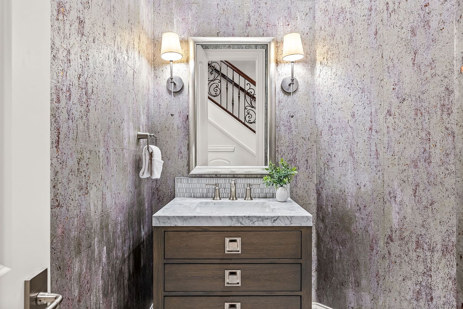 This is a well-decorated guest bathroom featuring a vanity with a marble countertop and a wooden cabinet with three drawers. A silver-framed mirror hangs above the sink, flanked by two sconce lights. The walls are covered in textured wallpaper, adding a touch of elegance to the space.