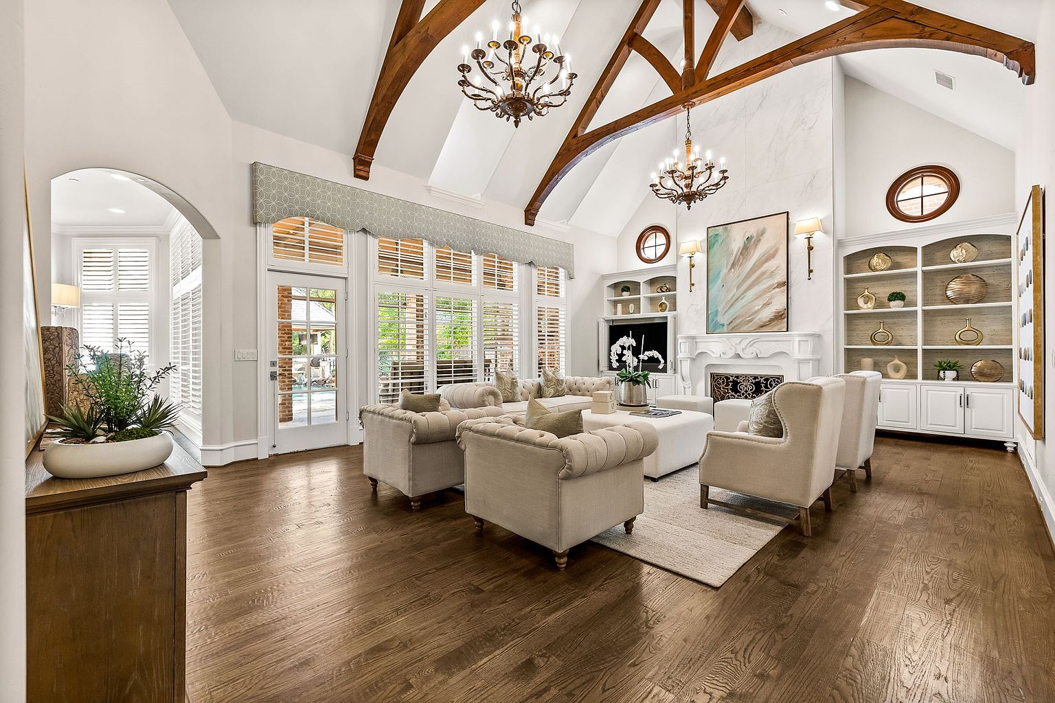 This is an interior shot of a luxurious living room featuring hardwood floors, exposed wooden beams, and elegant chandeliers. The room is furnished with comfortable, neutral-toned seating arranged around a light-colored rug and ottoman. Large windows with shutters provide ample natural light, and built-in shelving units flank a fireplace, adding to the room's sophisticated and inviting atmosphere.