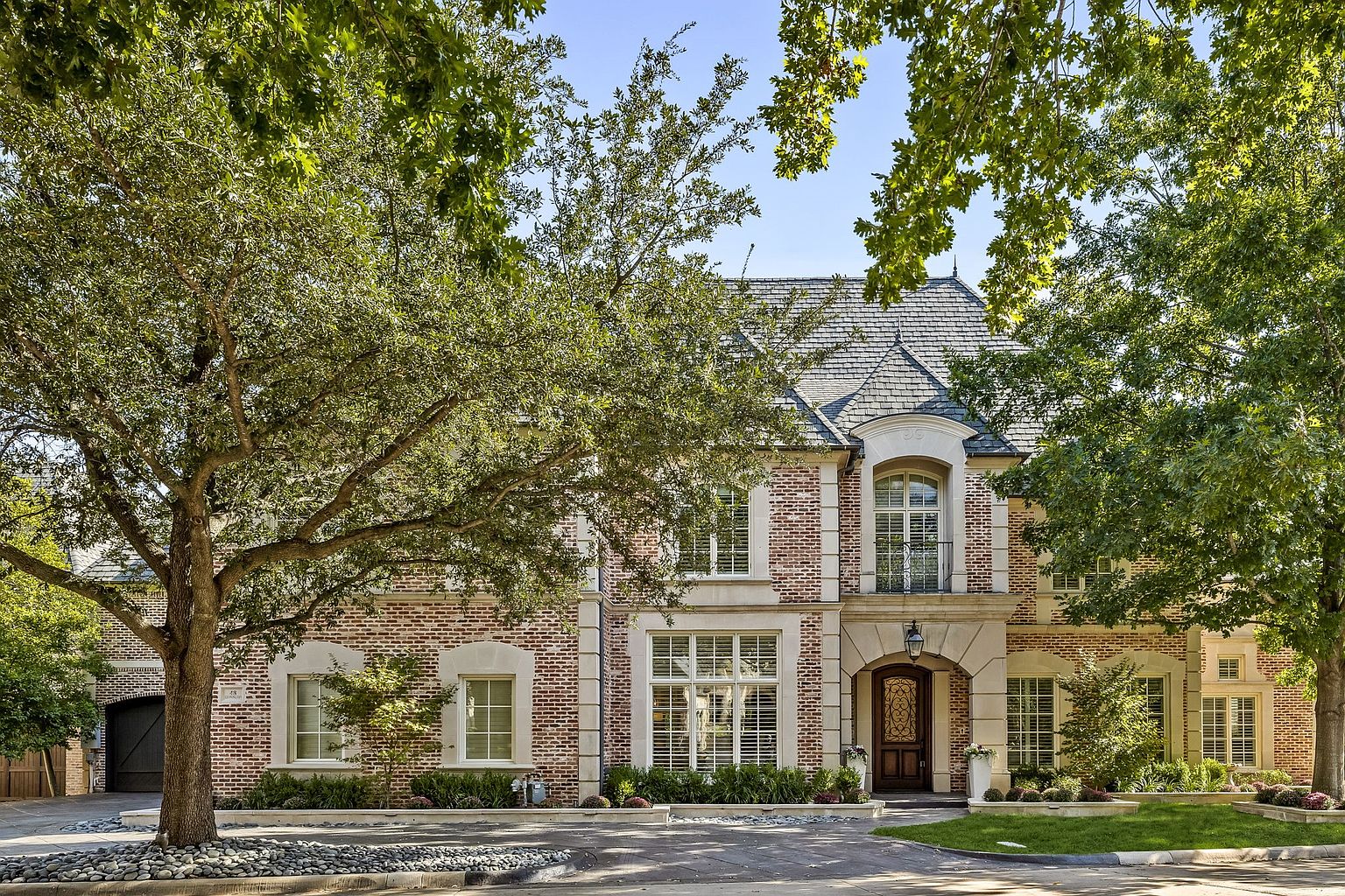 This is a front exterior view of a stately brick home with a gray slate roof. The architecture features arched windows, stone accents, and a prominent entryway with a wooden door. Mature trees frame the house, adding to its curb appeal and creating a sense of established elegance.