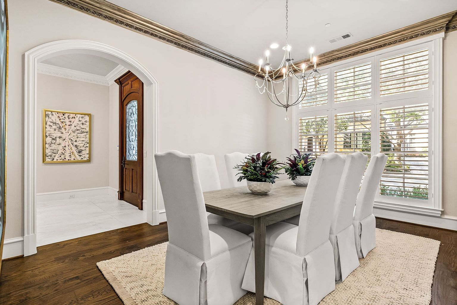 This is an interior shot of a dining room featuring a wooden table with white upholstered chairs. The room is well-lit by a chandelier and natural light coming through shuttered windows. A doorway leads to another room with a framed piece of art, and the flooring transitions from dark wood to light tile, adding visual interest.