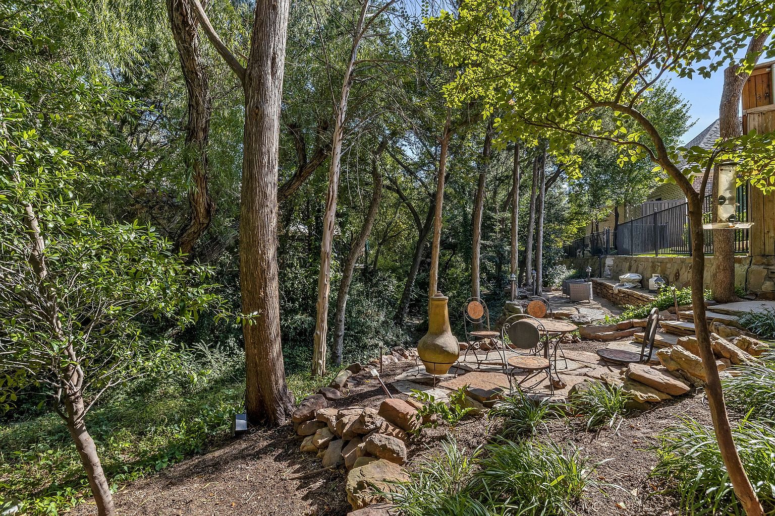 This image showcases a beautifully landscaped backyard garden area. A stone patio features a table and chairs, creating an inviting outdoor seating space. The garden is surrounded by mature trees and lush greenery, offering a sense of privacy and tranquility, making it an ideal spot for relaxation and entertainment.