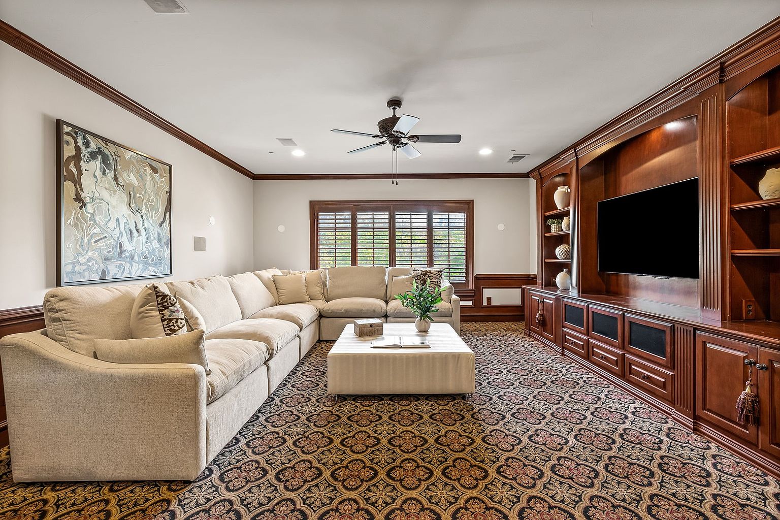 This is an interior shot of a living room featuring a large, comfortable sectional sofa and a custom-built entertainment center. The room is decorated with a patterned carpet, a neutral color palette, and a large piece of artwork. The space feels inviting and well-appointed, perfect for relaxation and entertainment.