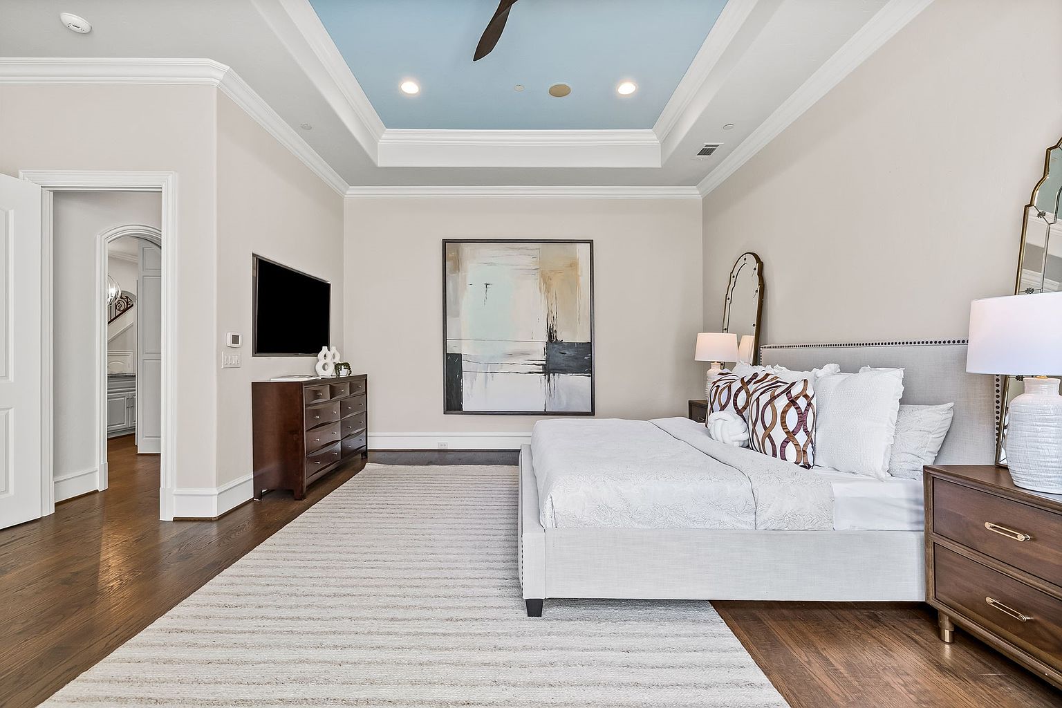 This is a well-lit primary bedroom featuring a large bed with white linens and decorative pillows, a dark wood dresser with a TV, and an abstract painting above the bed. The room has hardwood floors, a light-colored rug, and a tray ceiling with a blue accent. The perspective is from the doorway, showcasing the room's layout and design.