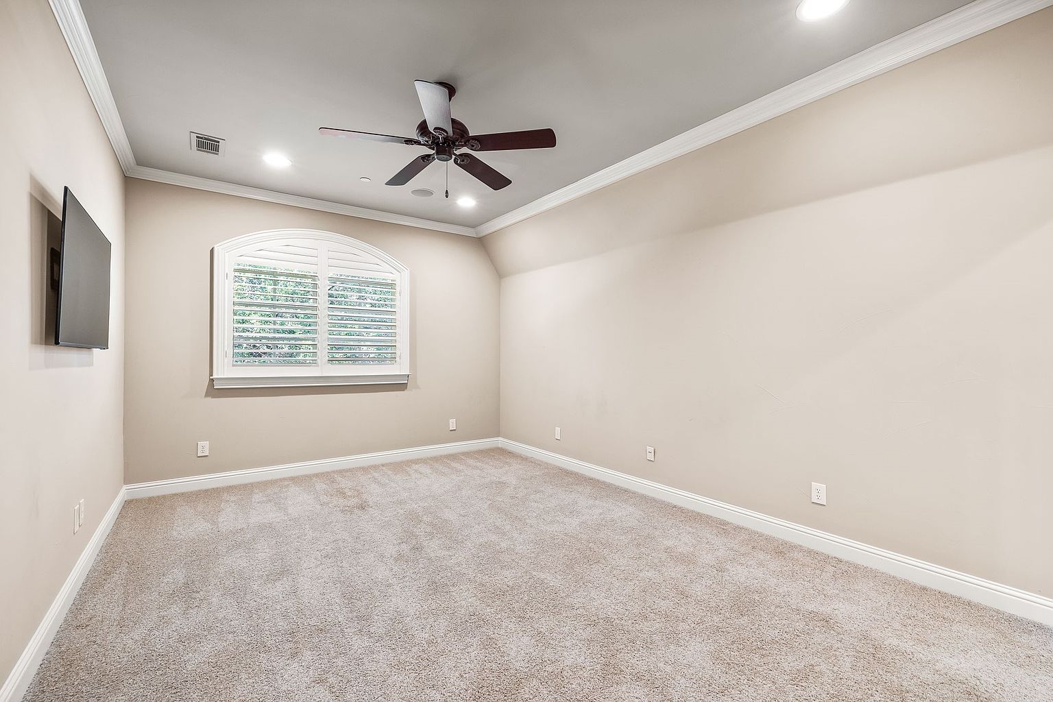 The image showcases a well-lit, empty bedroom with neutral beige walls and carpet. A ceiling fan with dark blades is centered in the room, and a window with white shutters provides natural light. A flat-screen television is mounted on the left wall, suggesting a space ready for personalization and comfort.