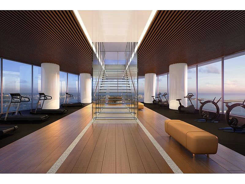 This is a modern home gym featuring wood floors with a white stripe down the center, floor-to-ceiling windows with a view, and a variety of exercise equipment including treadmills and stationary bikes. The gym also has white cylindrical pillars and a glass staircase in the center. The overall impression is clean, spacious, and luxurious.