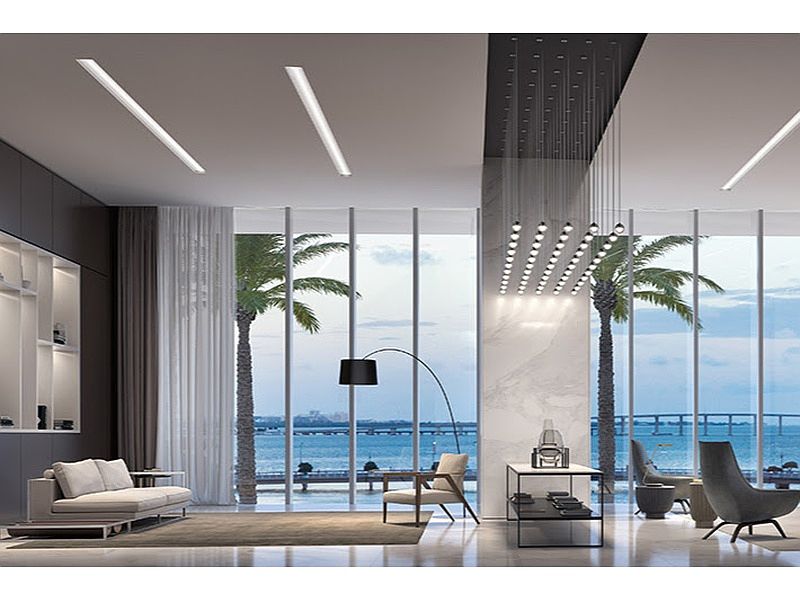This is a luxurious living room with floor-to-ceiling windows offering stunning ocean views framed by palm trees. The room features modern furniture, including a sleek sofa, stylish chairs, and a contemporary floor lamp. The overall design is minimalist and elegant, creating a serene and inviting atmosphere.
