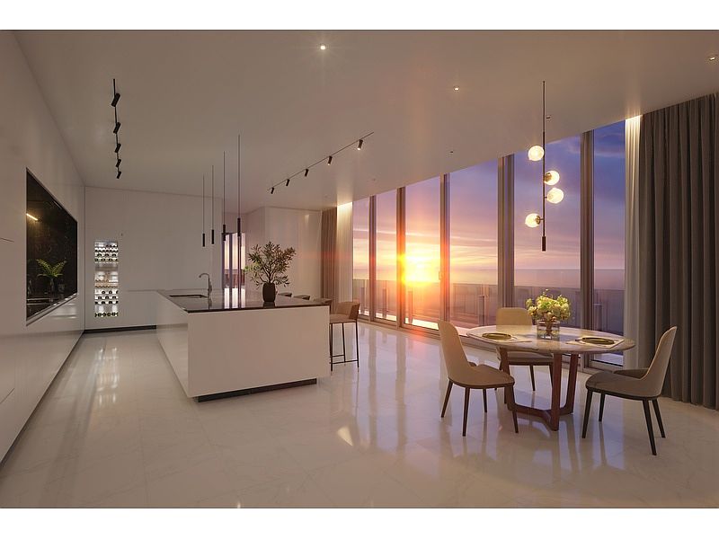 This is a modern, high-end kitchen and dining area featuring sleek white cabinetry, a large island with a dark countertop, and floor-to-ceiling windows offering a stunning sunset view. The dining area includes a round table with four chairs and a modern pendant light fixture, creating an elegant and inviting atmosphere. The perspective is wide, showcasing the open-concept design and luxurious finishes.