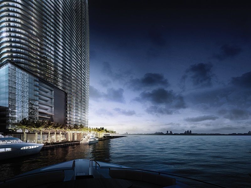 This image showcases a modern high-rise building from a waterfront perspective, emphasizing its architectural grandeur and luxurious waterfront access. Several yachts are docked along the pier, highlighting the upscale lifestyle associated with the property. The dramatic sky and city skyline in the distance add depth and a sense of location.