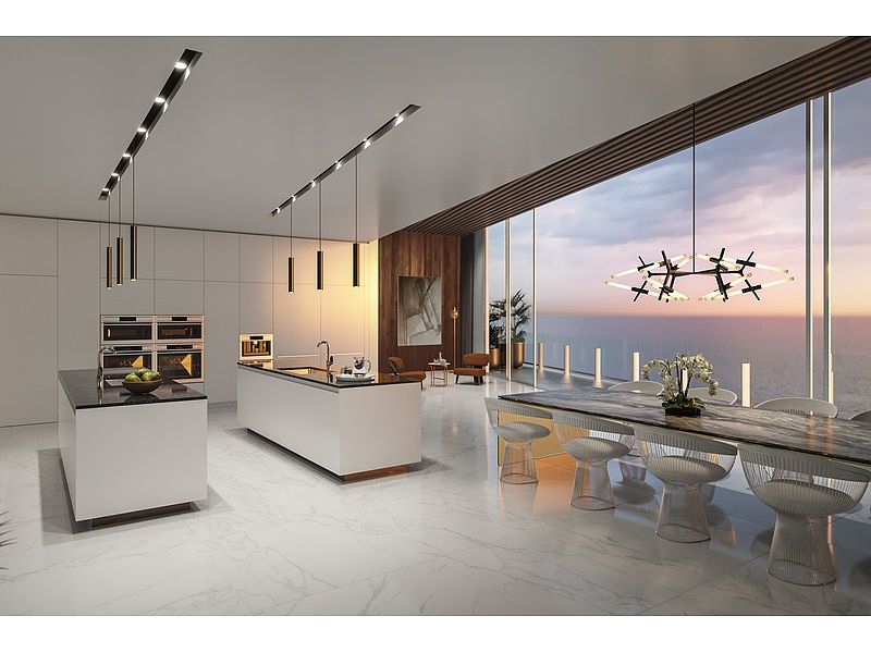 This is a modern, luxury kitchen with sleek white cabinetry, a large island with a dark countertop, and high-end appliances. The open-concept design flows into a dining area with floor-to-ceiling windows offering stunning ocean views. The space is illuminated by contemporary linear and geometric chandeliers, creating a sophisticated and inviting atmosphere.